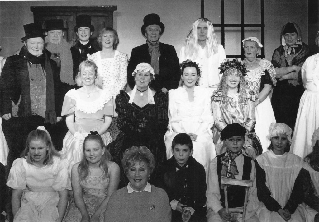 2002 A Christmas Carol | Back Row: John Chivers, Jim Biggins, James Wakefield, Fran Creasey, John Lawrence, Clive Heginbotham, Enid Taylor, Sue Heginbotham. Middle Row: Stephanie Milburn, Barbara Colbourne, Charlotte Wakefield, Avril Allsop, Karen South. Front row: Christine Lawrence and children.