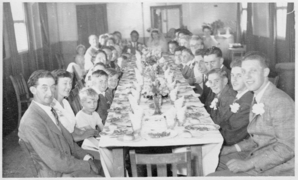 1954. Old village hall interior at wedding reception of Les Matthews & Dorothy Milton who are sat facing camera at far end of table. From front, Bill Godwin is 1st on left, Derrick Godwin 4th on left, Colin Godwin 5th on left. Roger Weston 1st on right, Roy Godwin 2nd right, John ‘Pixie’ Miller 3rd right, Jim Taylor 4th right, Graham Maslen 5th right, Flo Taylor 6th right. At the far (east) end is the kitchen, the entrance and WCs were at the west end.